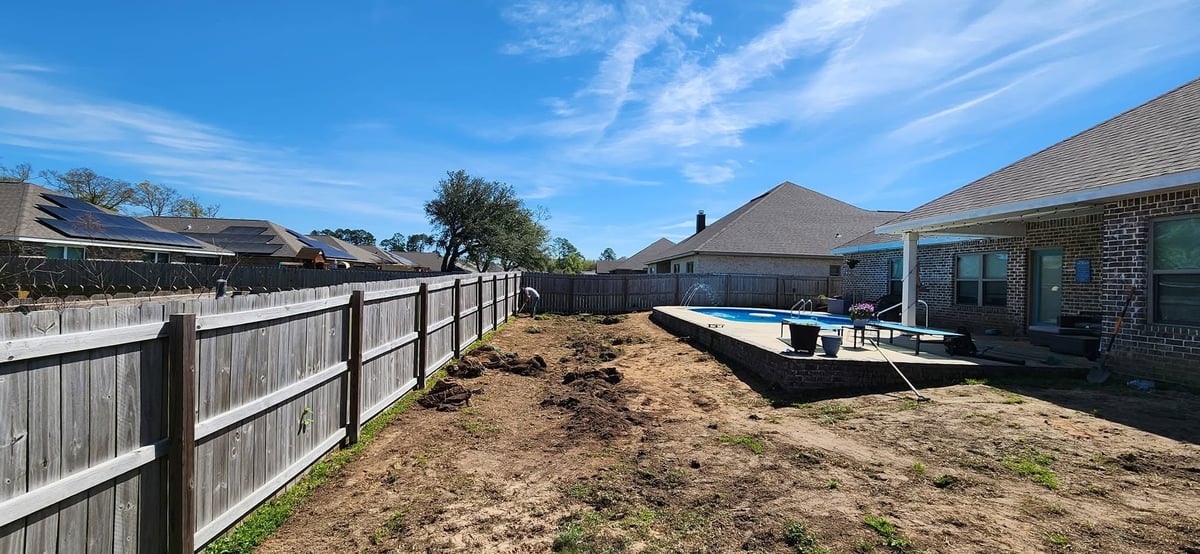 Backyard with wooden fence, bare ground, and blue sky, with houses and pool visible in background