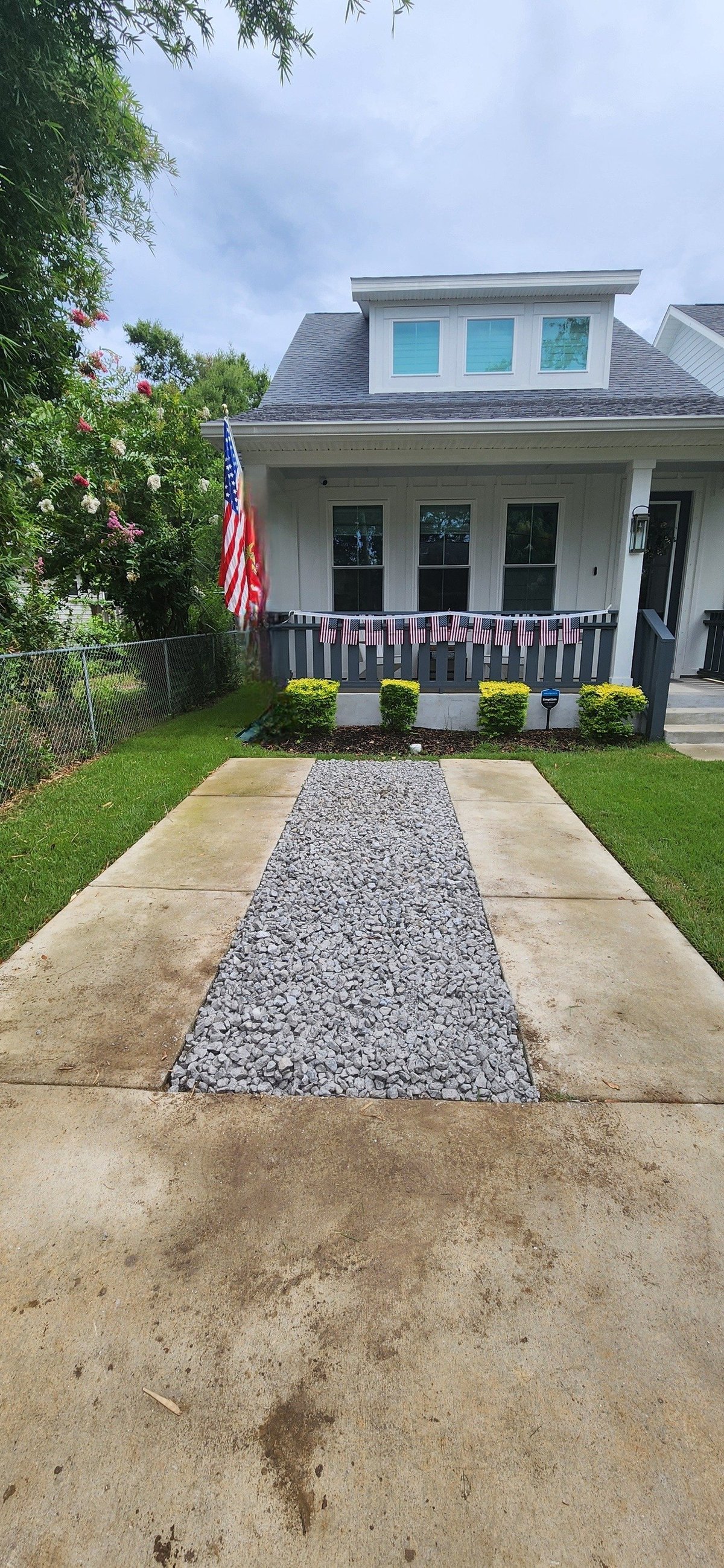 White colonial-style house with gray roof, front porch decorated with American flag, gravel pathway, and yellow landscaping shrubs