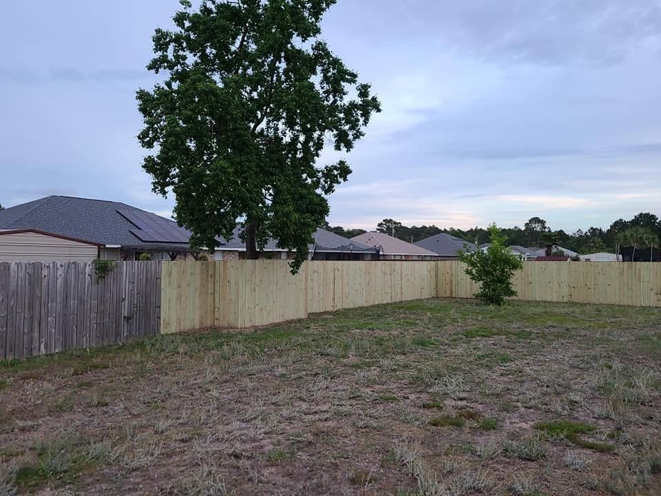 Empty backyard with wooden fence, large tree, and residential houses in background under cloudy sky