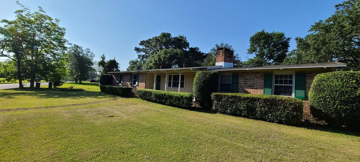 Single-story brick ranch house with green hedges on expansive lawn surrounded by mature trees under clear sky