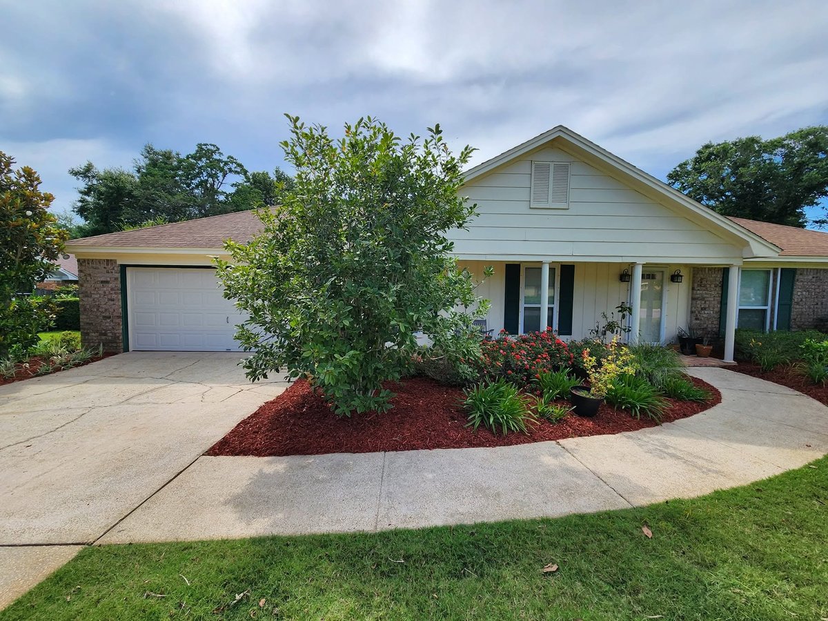 Single-story brick and siding home with white garage door, concrete driveway, landscaped front yard with red mulch and green plants under cloudy sky