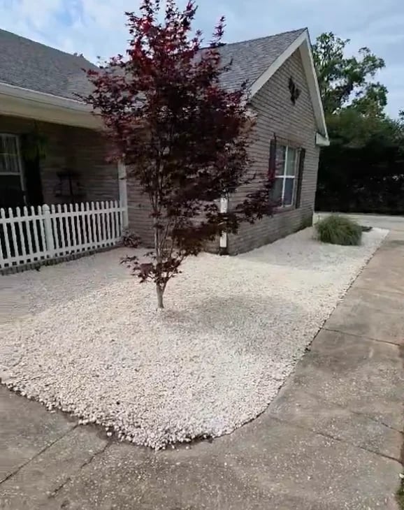Single-story house with white porch, burgundy tree in gravel yard, and concrete driveway