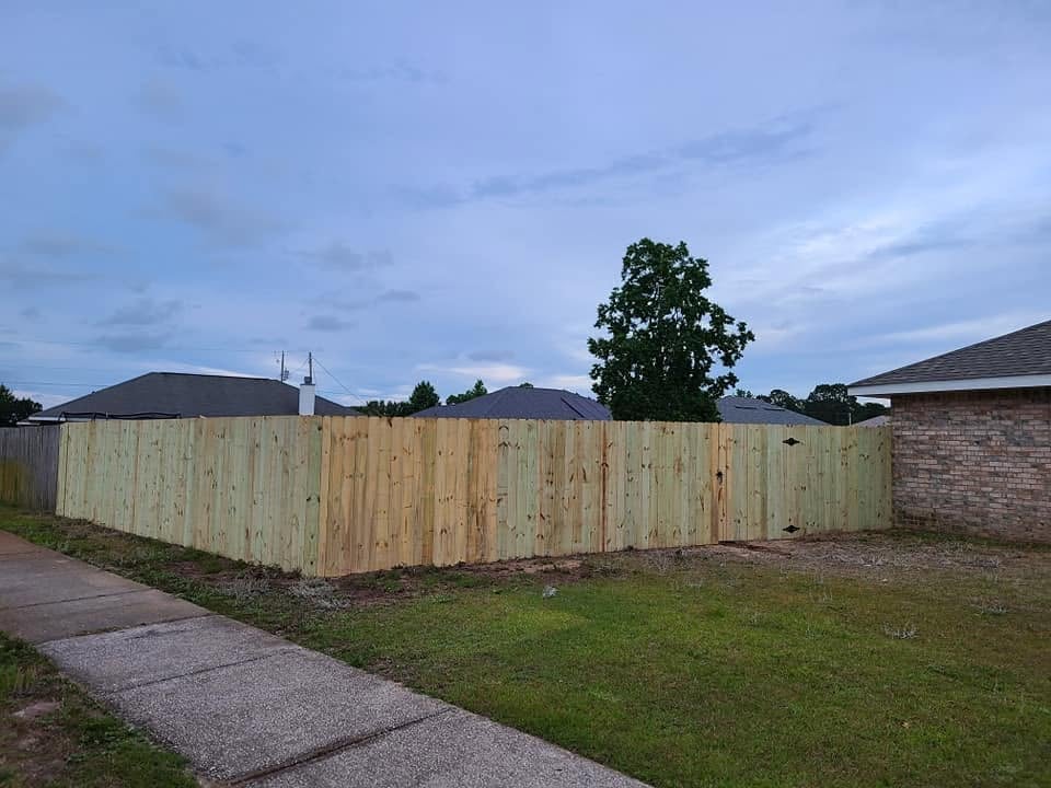 Suburban backyard with wooden fence, green lawn, concrete pathway, and trees under a cloudy blue sky