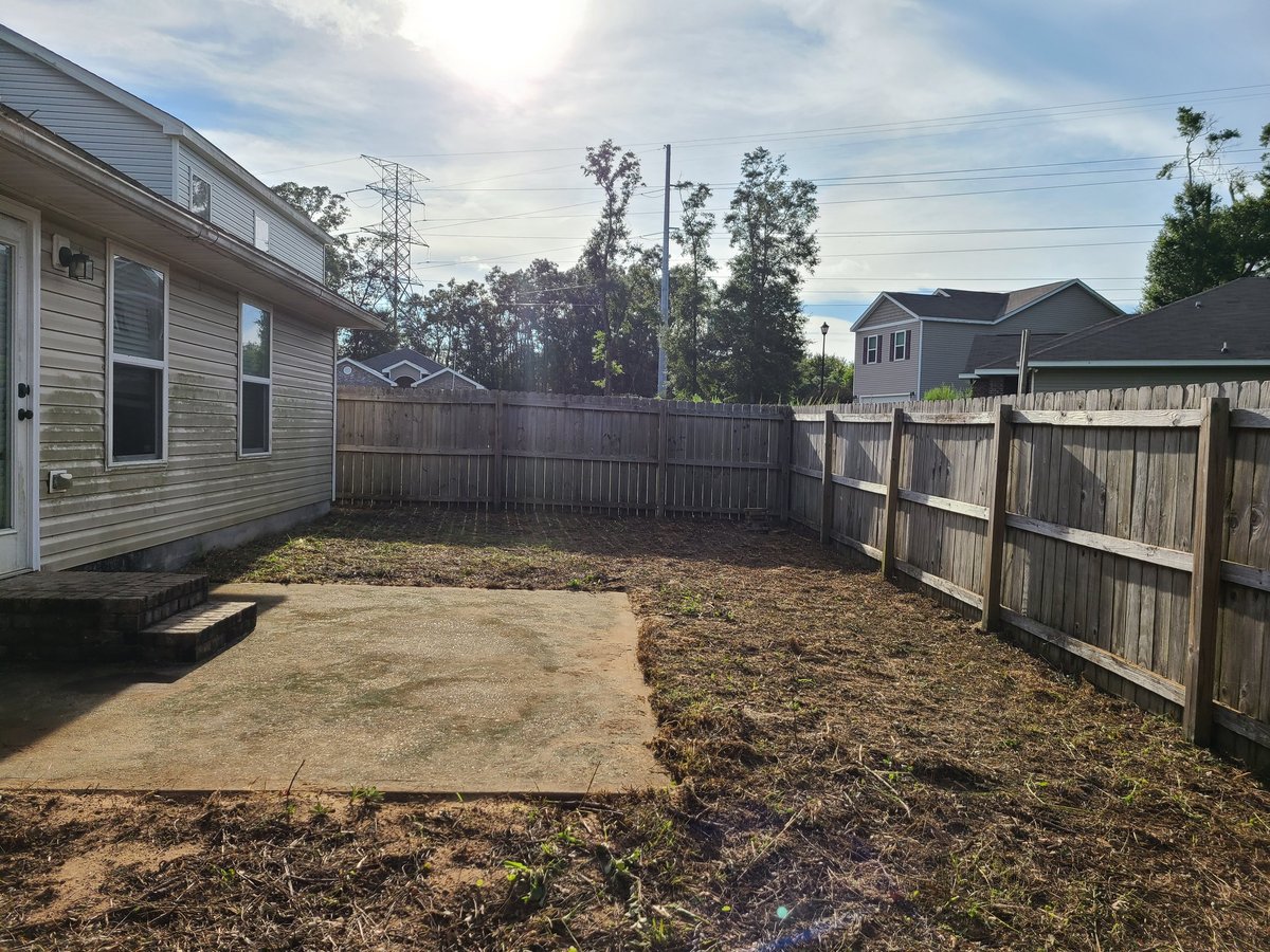 Residential backyard with wooden fence, bare ground, and adjacent houses under partly cloudy sky