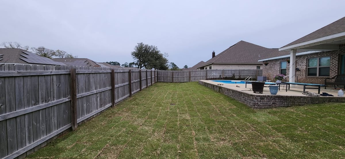 Spacious backyard with wooden fence, manicured lawn, patio with pool, and brick house on overcast day