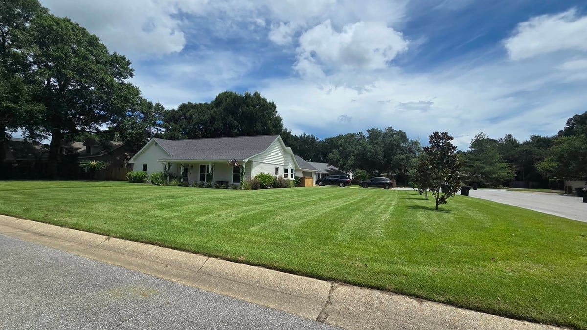 White single-story house with covered porch on a large manicured lawn with trees under partly cloudy sky